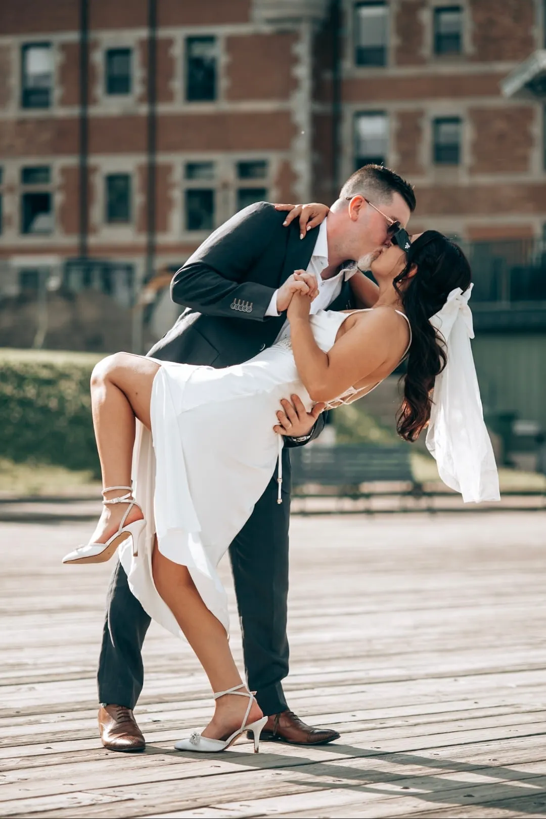 A couple kissing in a deep dip on a boardwalk, brick buildings behind them