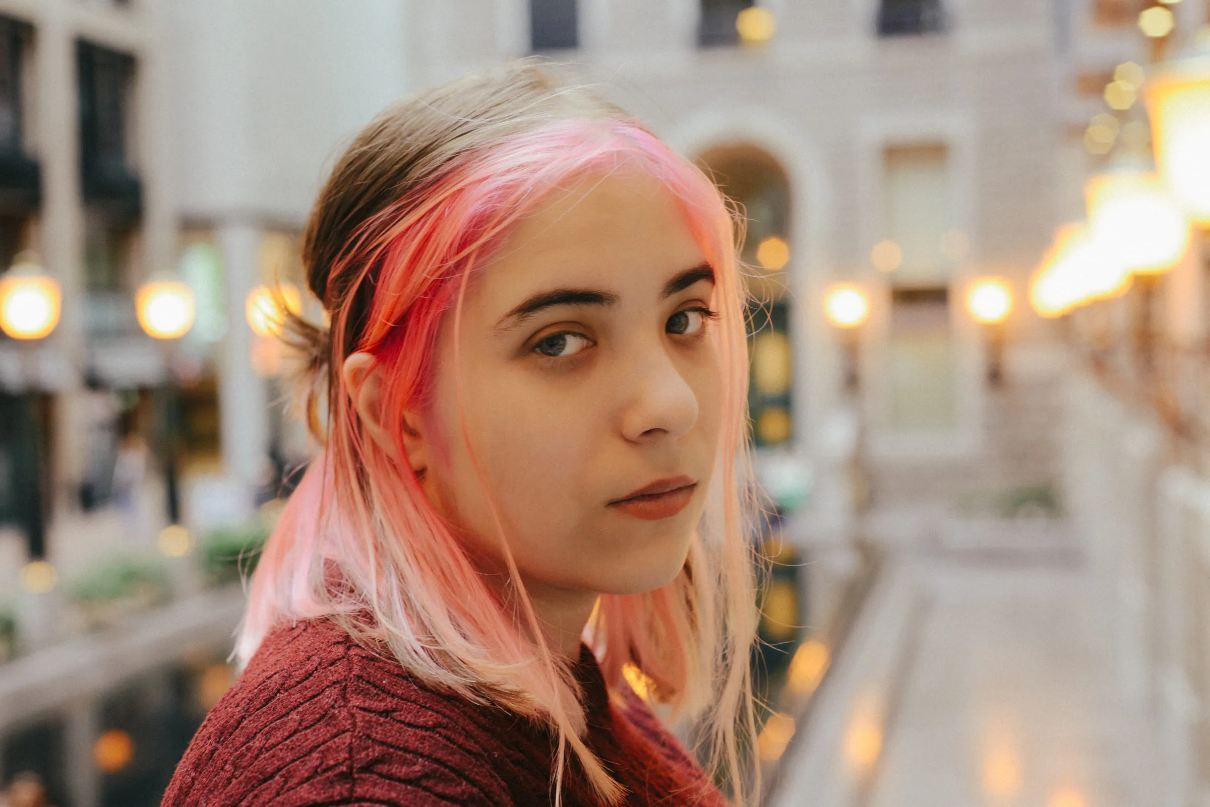 A portrait of a young woman with pink hair, warm city lights blurred behind her
