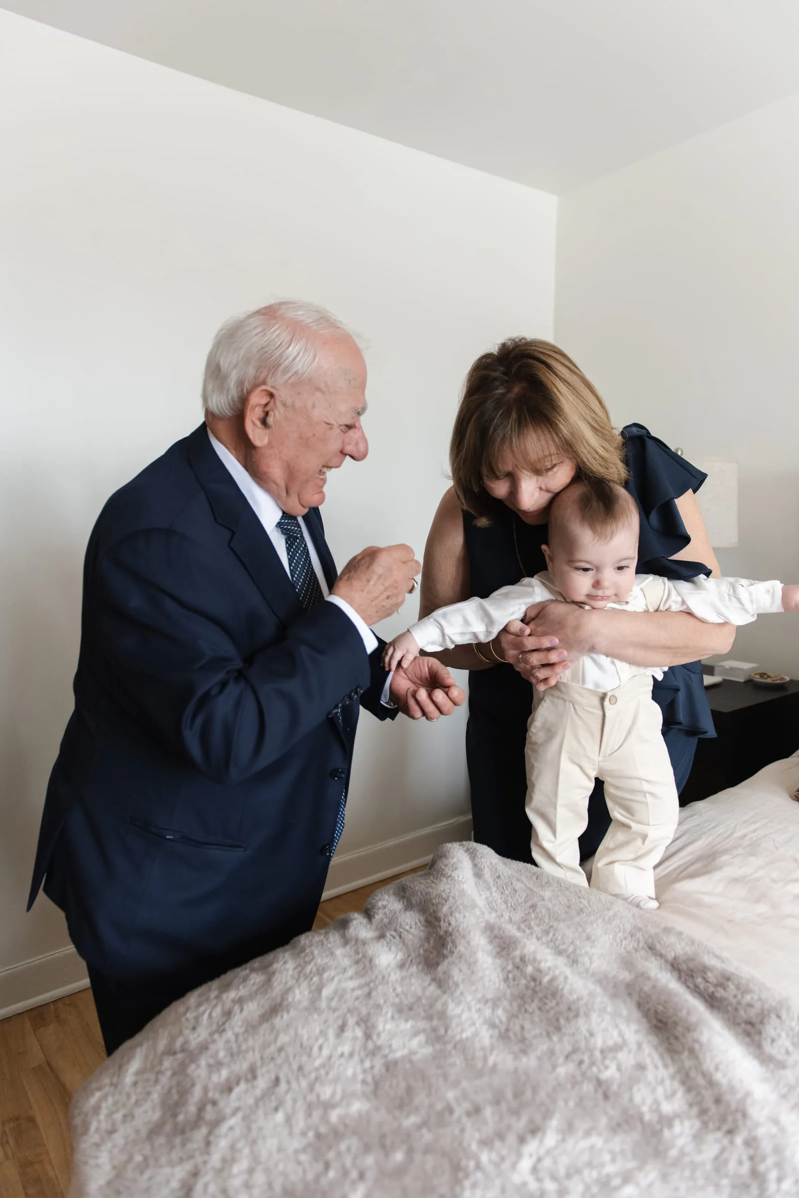 An older couple holding the baby’s hands as he stands on the bed