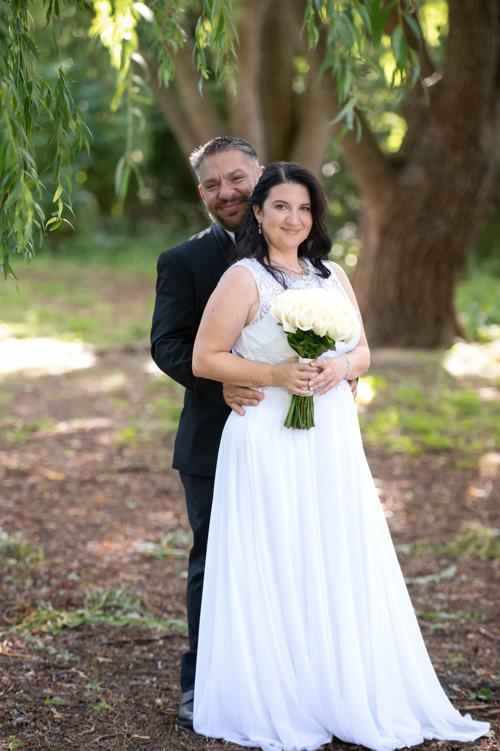 The bride and groom standing close together in dappled light beneath a willow tree