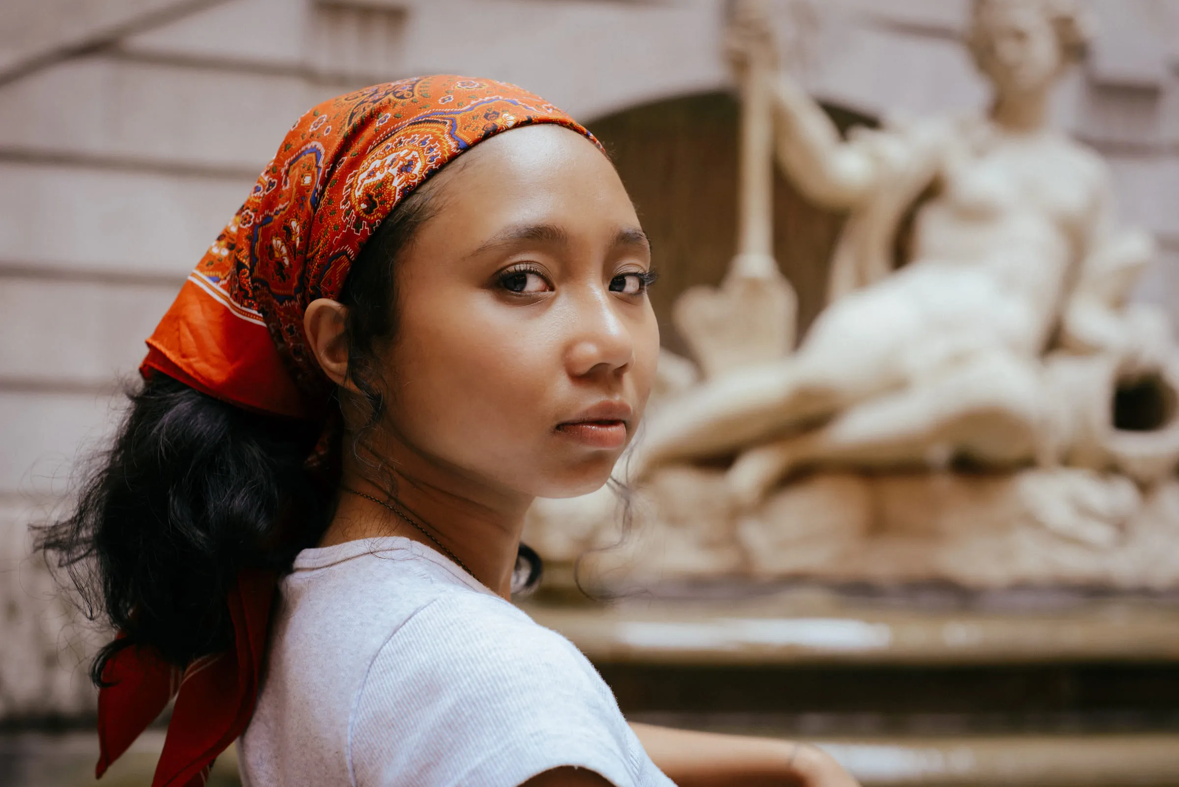 A young woman in an orange headscarf looking back at the camera in front of a carved stone sculpture