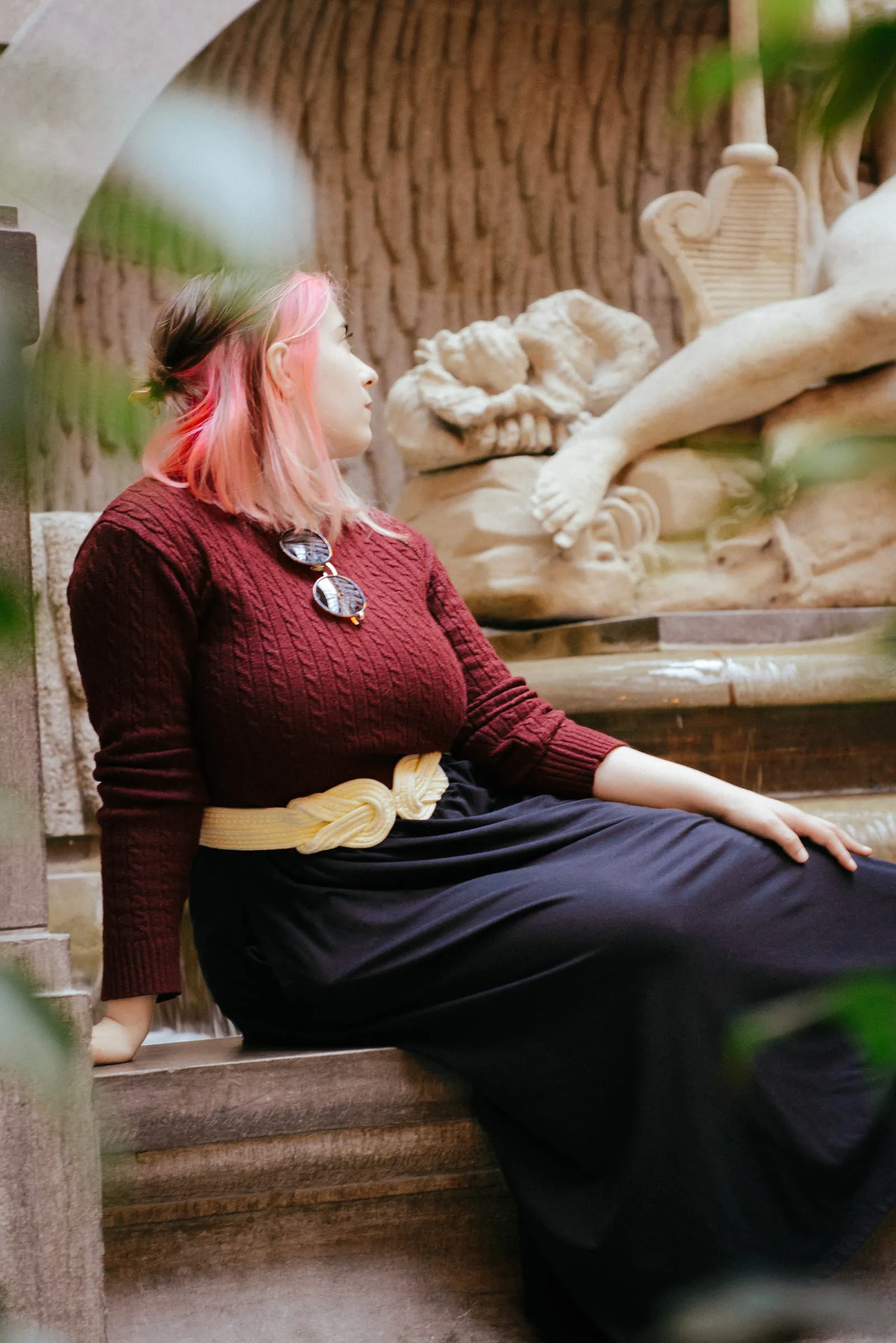 A young woman seated against a stone ledge below a reclining sculpture