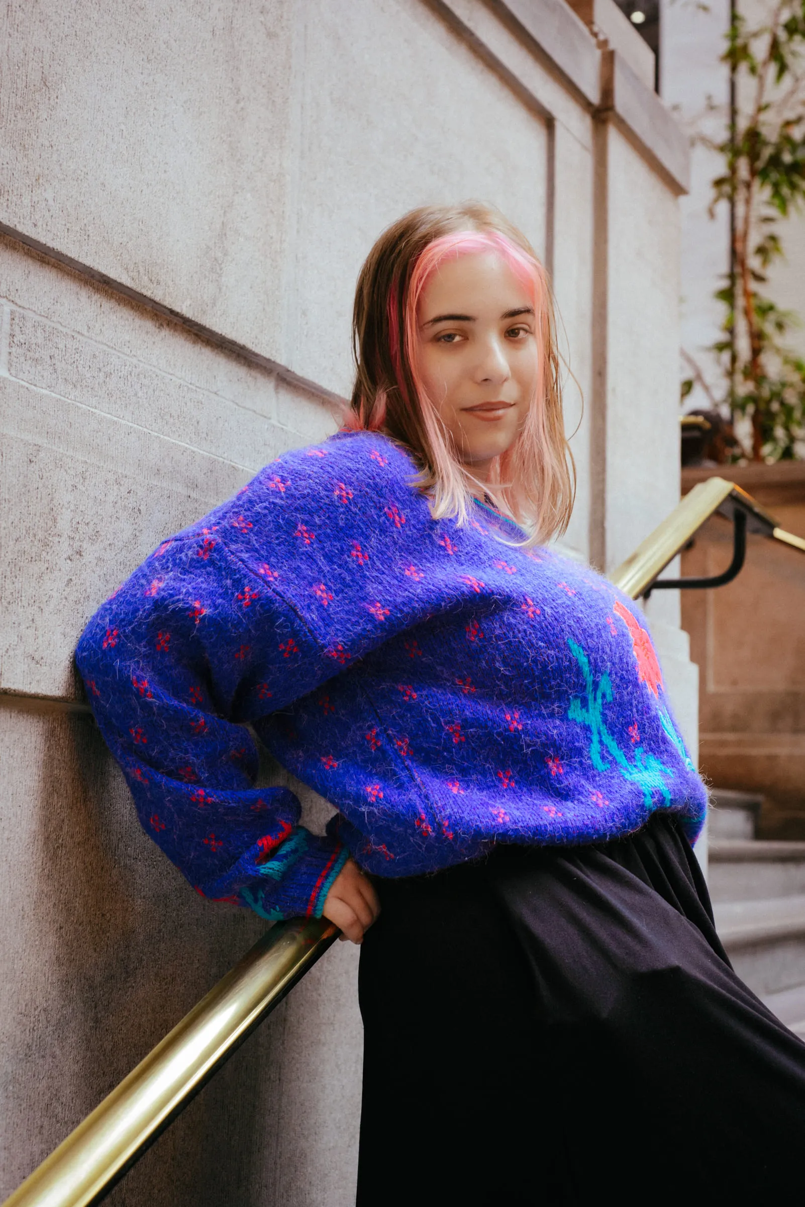 A young woman in a bright blue mohair sweater leaning against a brass handrail