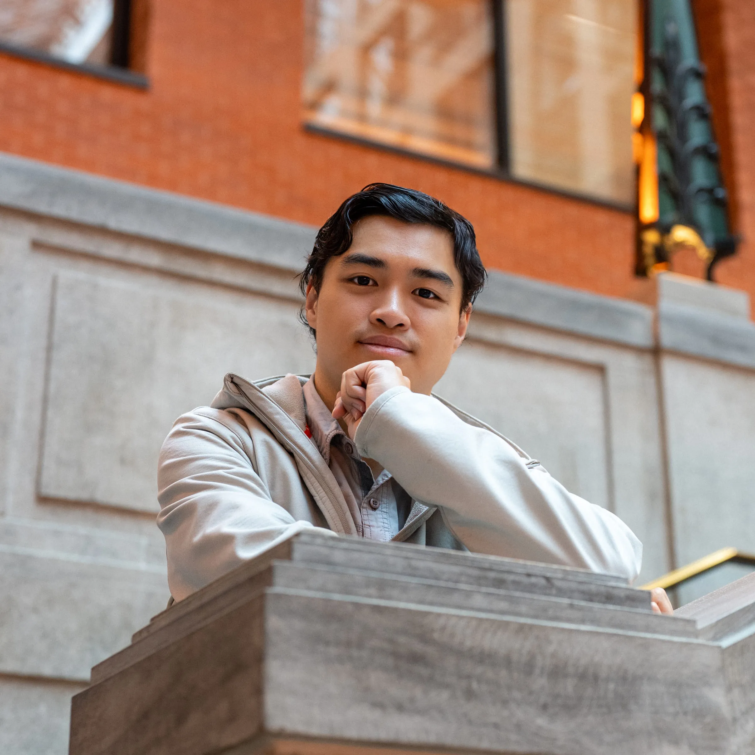 A young man resting his chin on his hand against a stone ledge, red brick behind him