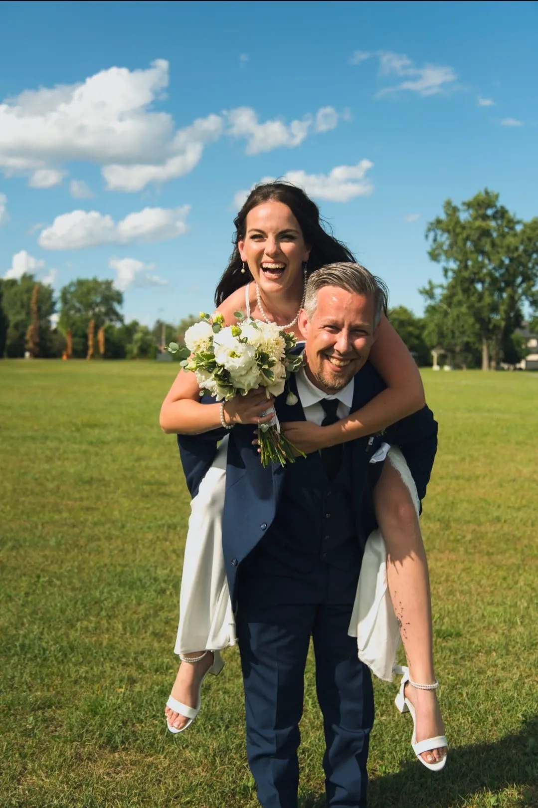 A groom carrying his bride on his back across a sunlit lawn, both of them laughing