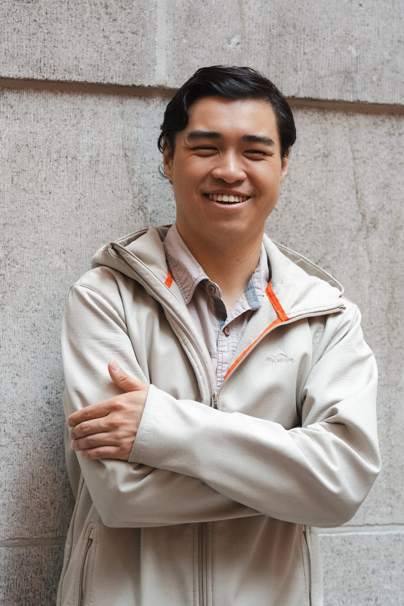 A young man laughing with his arms folded, against a stone wall