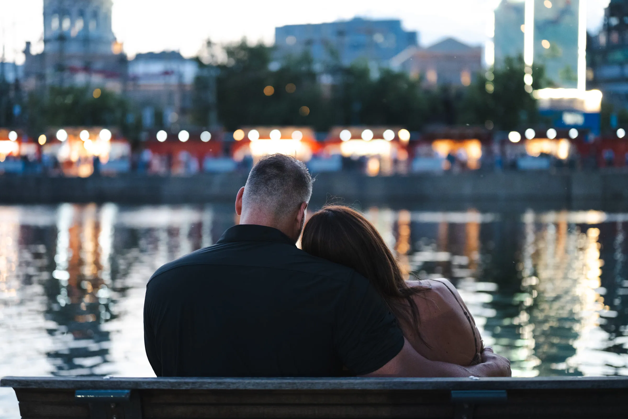 The two of them on a bench, heads together, facing the lights across the water