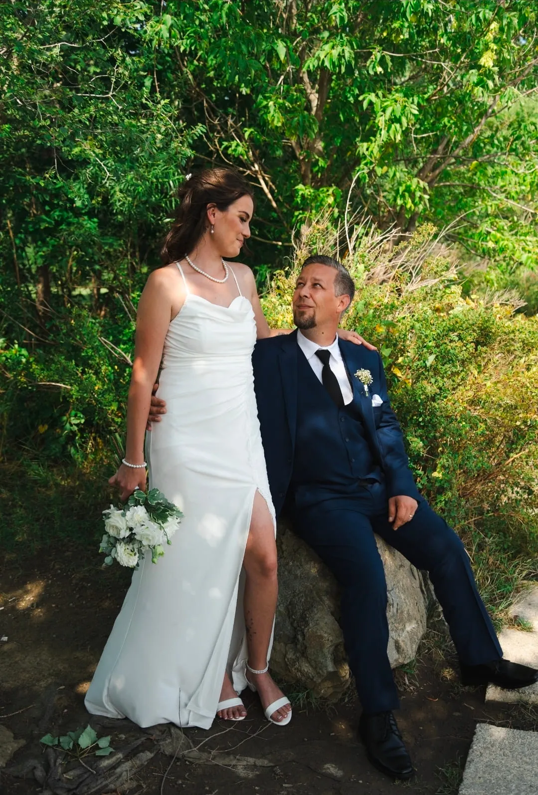 A bride standing beside her groom, who is seated on a rock under summer trees