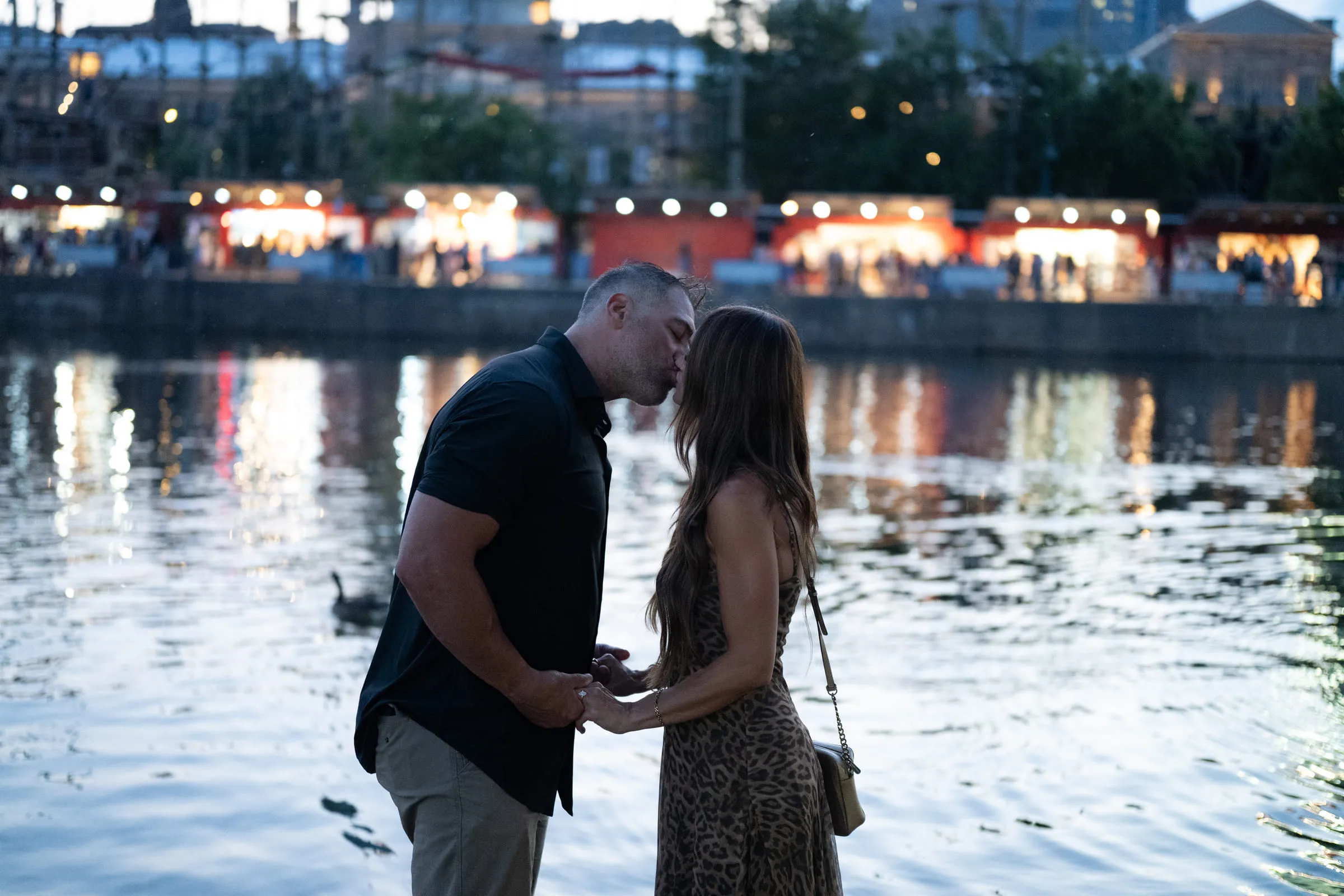 The couple at the water’s edge as the harbour lights come on behind them