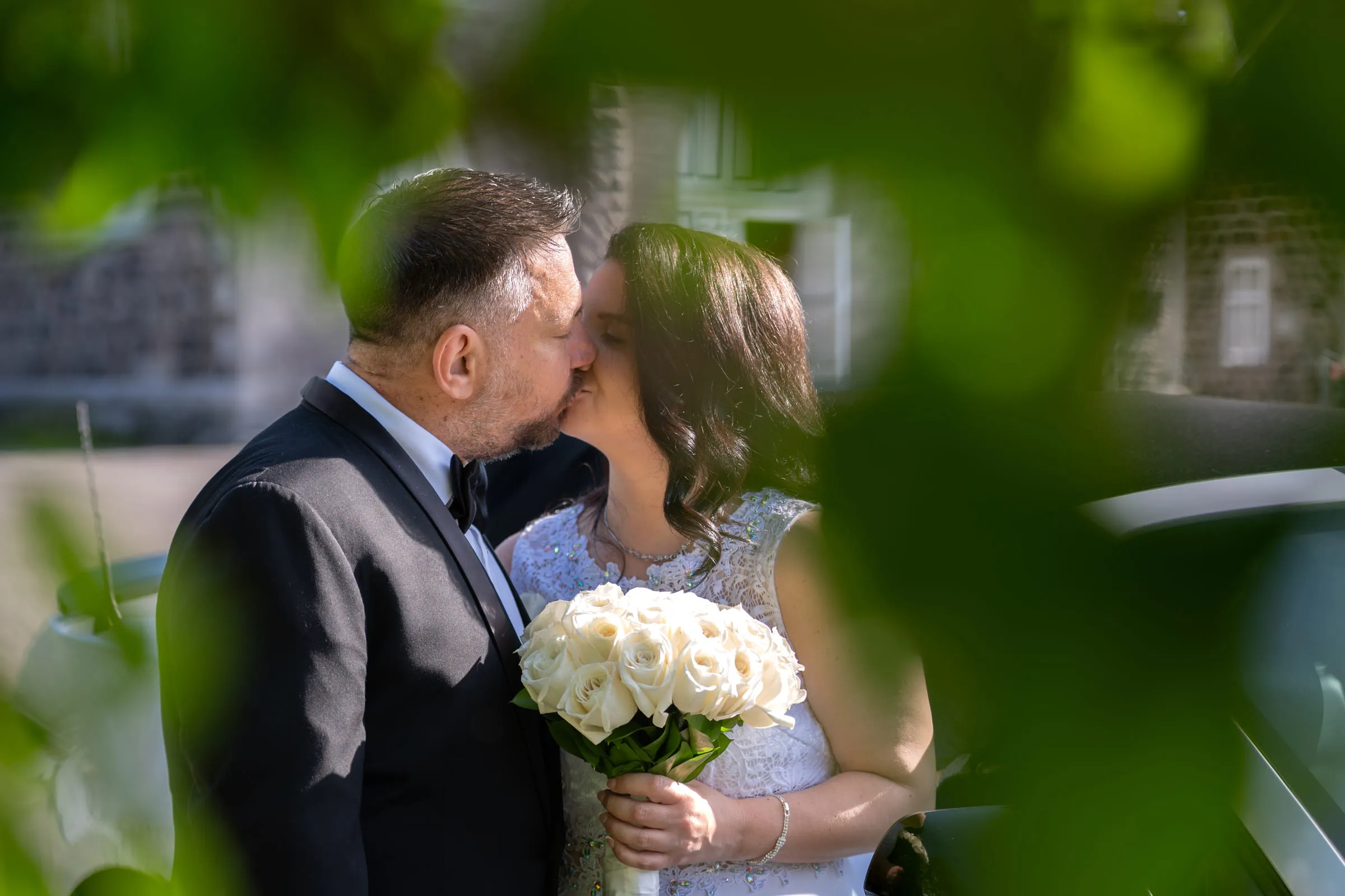 A bride and groom kissing, framed by out-of-focus green leaves